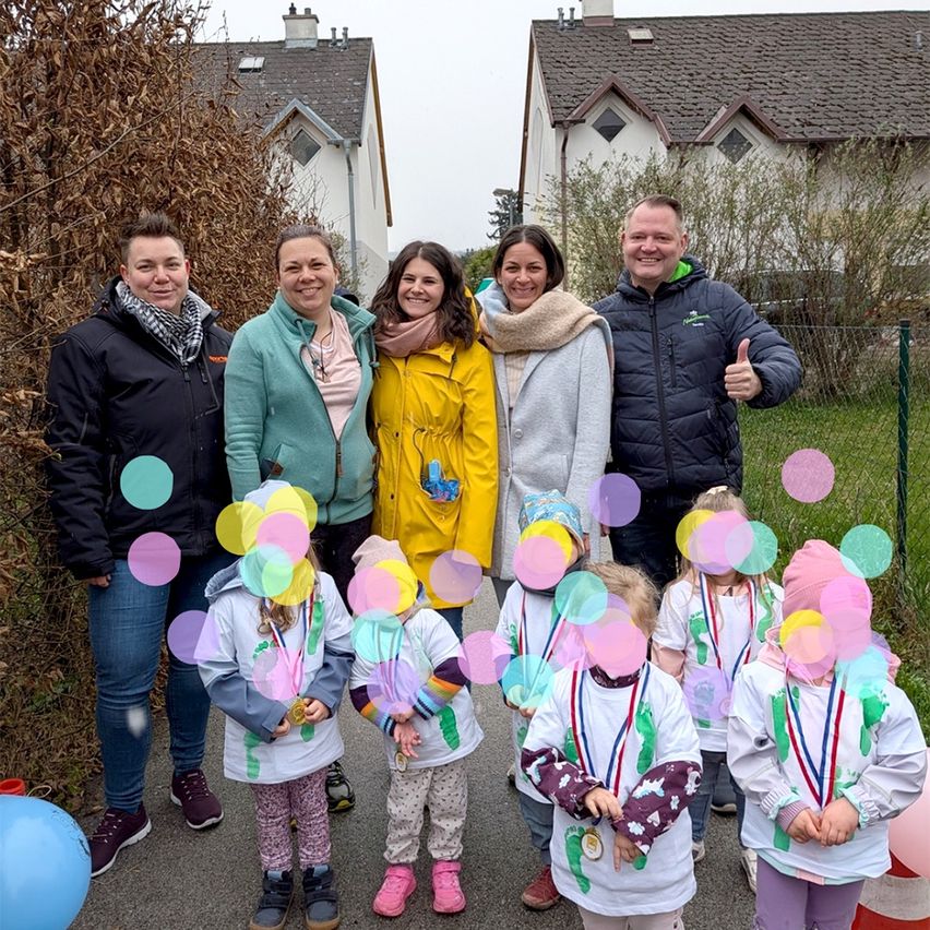 Eine Gruppe von Erwachsenen und Kindern posiert für ein Foto im Freien. Die Kinder tragen Medaillen und die Erwachsenen lächeln. Sie stehen vor einem Haus mit einem Zaun und grünem Rasen.