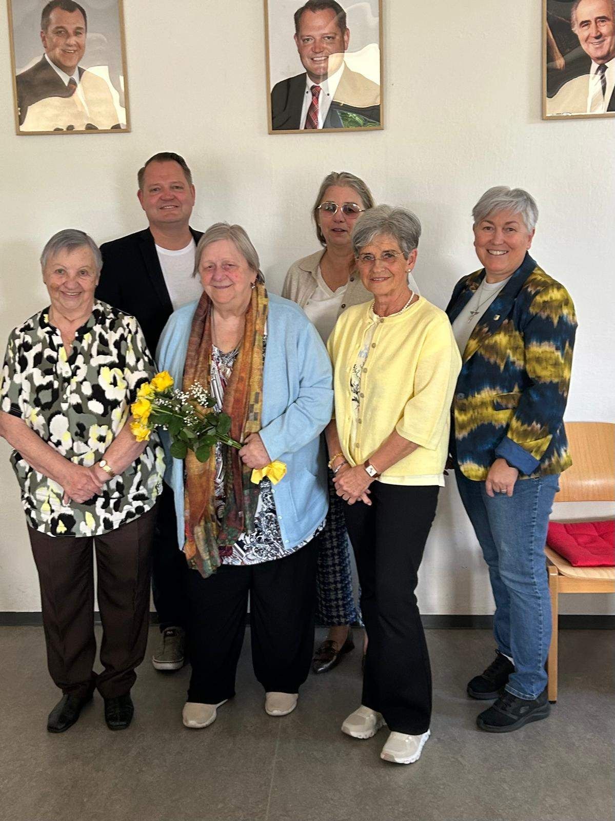 A group of women and men stand together in a room with a white wall. A woman holds flowers.