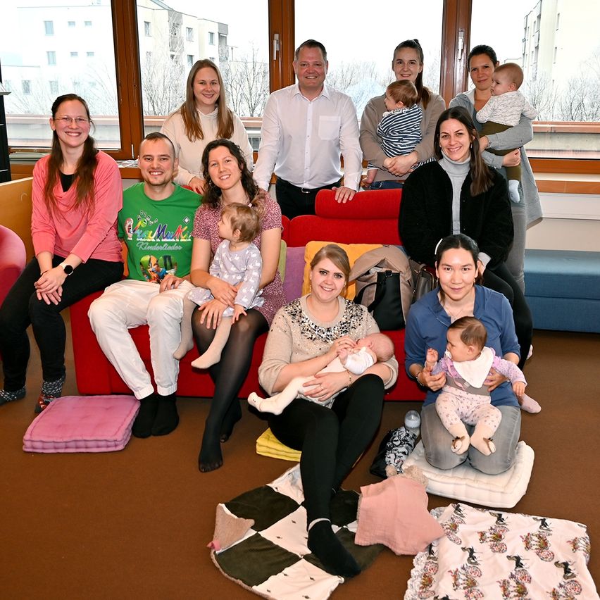 Eine Gruppe von Erwachsenen und Kindern posiert für ein Foto in einem Raum mit einem roten Sofa und Kissen, einige halten Babys.