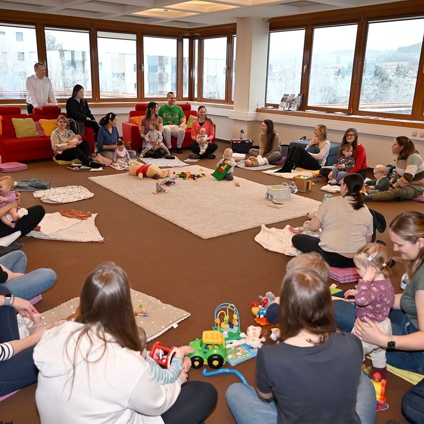 Eine Gruppe von Erwachsenen und Kindern sitzt in einem Kreis in einem Raum mit Spielzeug auf dem Boden. Ein Mann steht in der Nähe des Fensters.