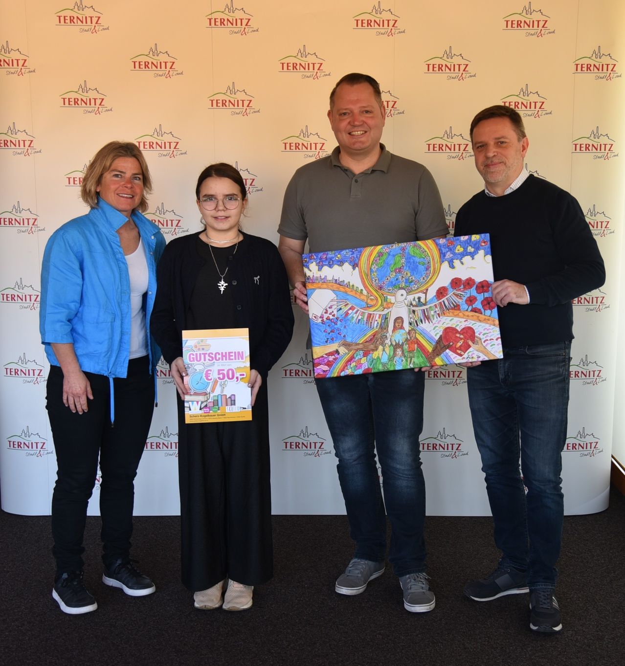 Four people stand in front of a wall with the word Ternitz. Two men hold a colorful painting and a book with a gift certificate. The woman in glasses holds a book. They all smile.