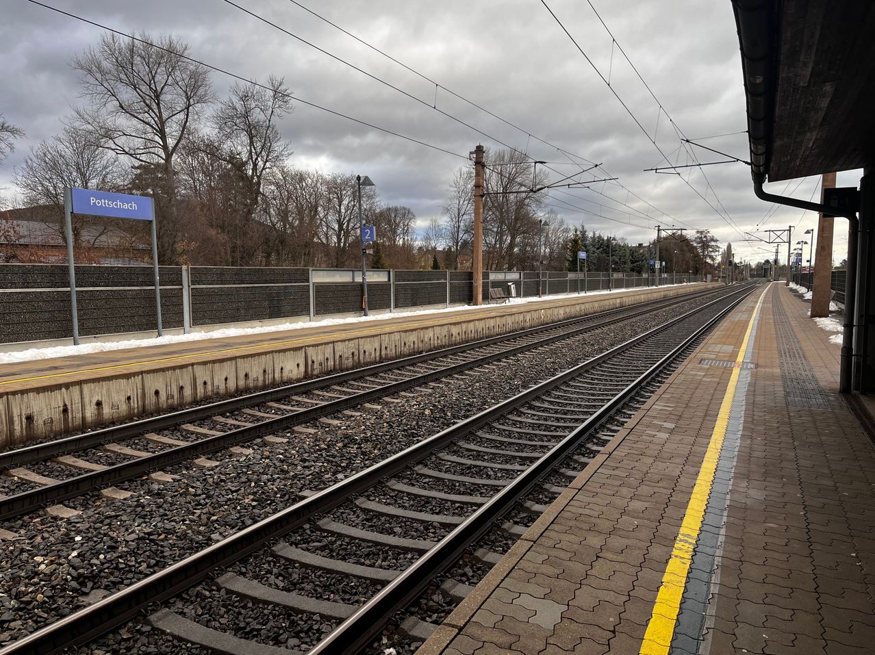 Ein leerer Bahnhof mit zwei Gleisen und Schotter an den Seiten. Der Bahnhof hat ein blaues Schild mit dem Wort Eschach. Bäume stehen auf der anderen Seite des Zauns.