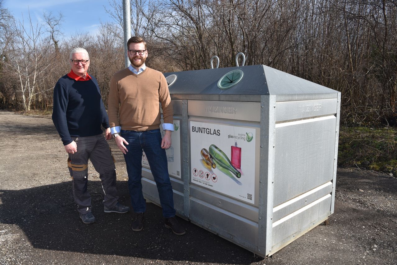 Zwei Männer stehen neben einem Buntglas-Recyclingbehälter mit einem grünen Kreis oben und einer Werbung an der Seite.