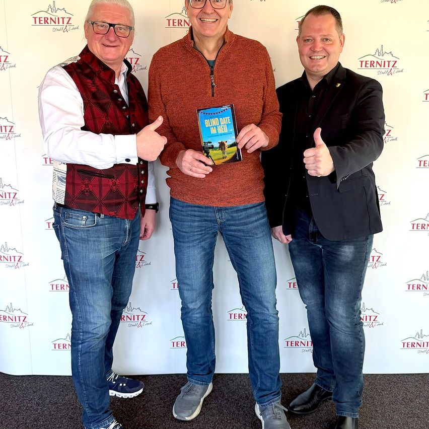 Drei Männer stehen zusammen, einer hält ein Buch mit dem Titel 'Blind Date in der Hölle', lächeln und geben Daumen hoch vor einer Wand mit mehreren 'TERNITZ' Logos.