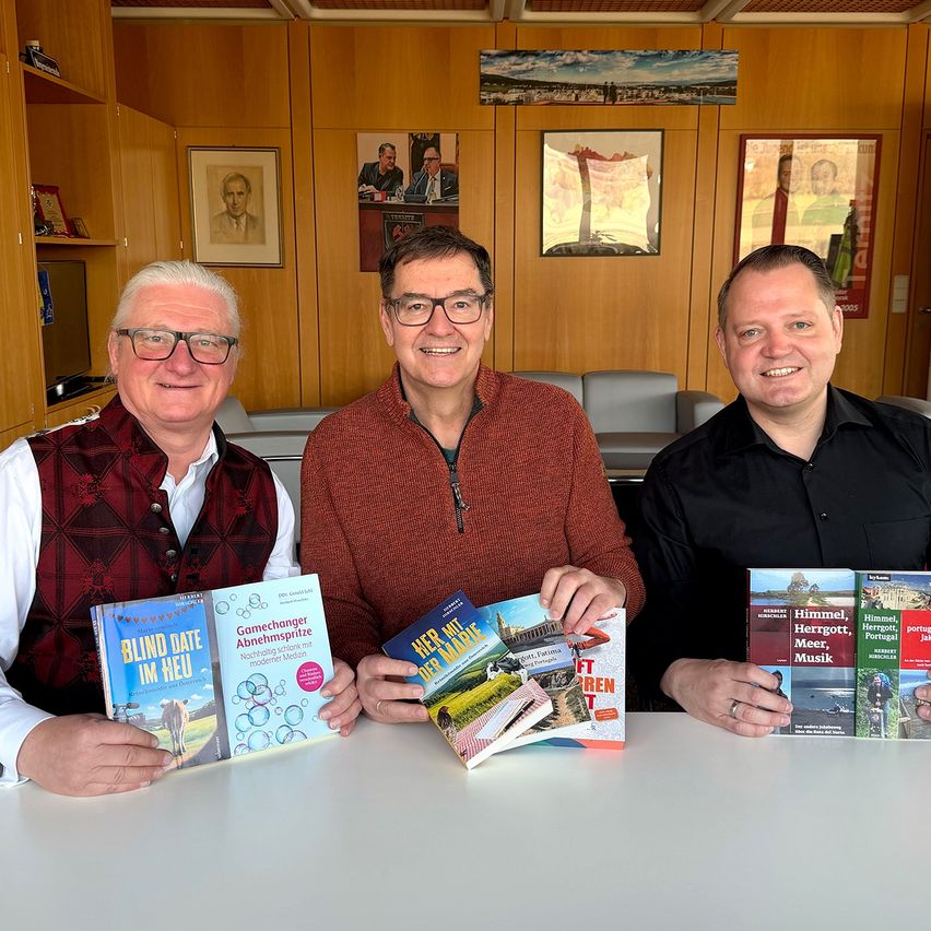 Drei Männer sitzen lächelnd an einem Tisch und halten mehrere Bücher in die Höhe. Der Mann auf der linken Seite hält ein Buch mit dem Titel Blind Date Im Neu. Der Mann in der Mitte hält ein Buch mit dem Titel Her Mit Der Nahe. Der Mann auf der rechten Seite hält mehrere Bücher. Sie sitzen vor einer Holzwand mit gerahmten Bildern und einer Tür.