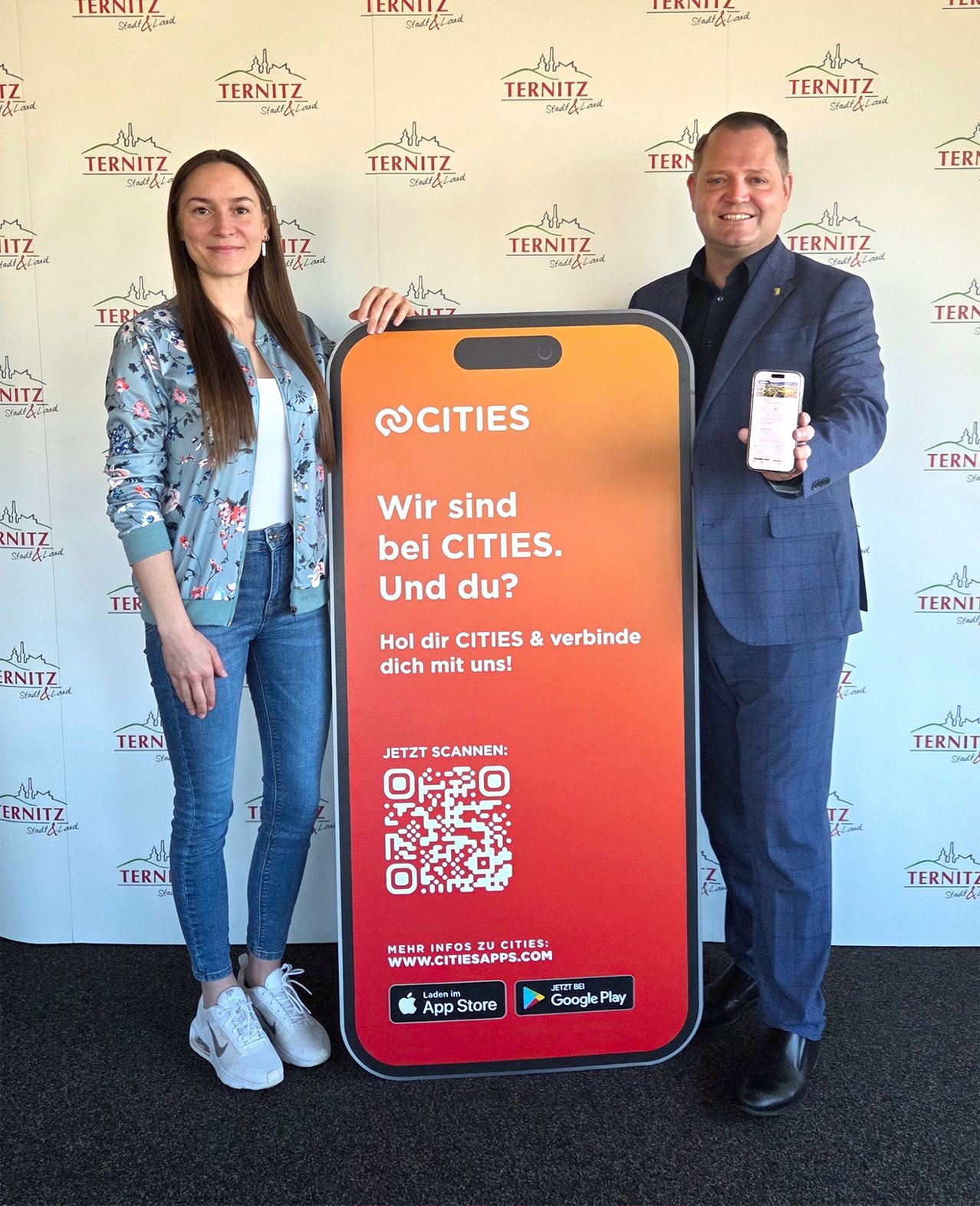 Ein Mann und eine Frau stehen neben einem Schild für die Cities-App und halten ein Smartphone. Das Schild zeigt 'Wir sind bei Cities. Und du?' und einen QR-Code. Der Hintergrund zeigt eine Wand mit dem Cities-Logo.
