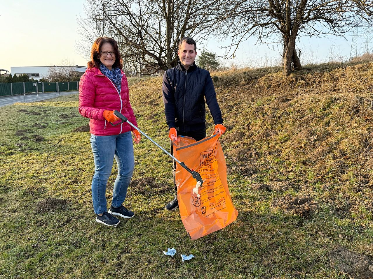 Ein Mann und eine Frau stehen auf einem Grasfeld und halten einen Müllsack. Die Frau trägt eine rosa Jacke und orange Handschuhe, der Mann eine blaue Jacke und schwarze Hose. Sie lächeln beide.