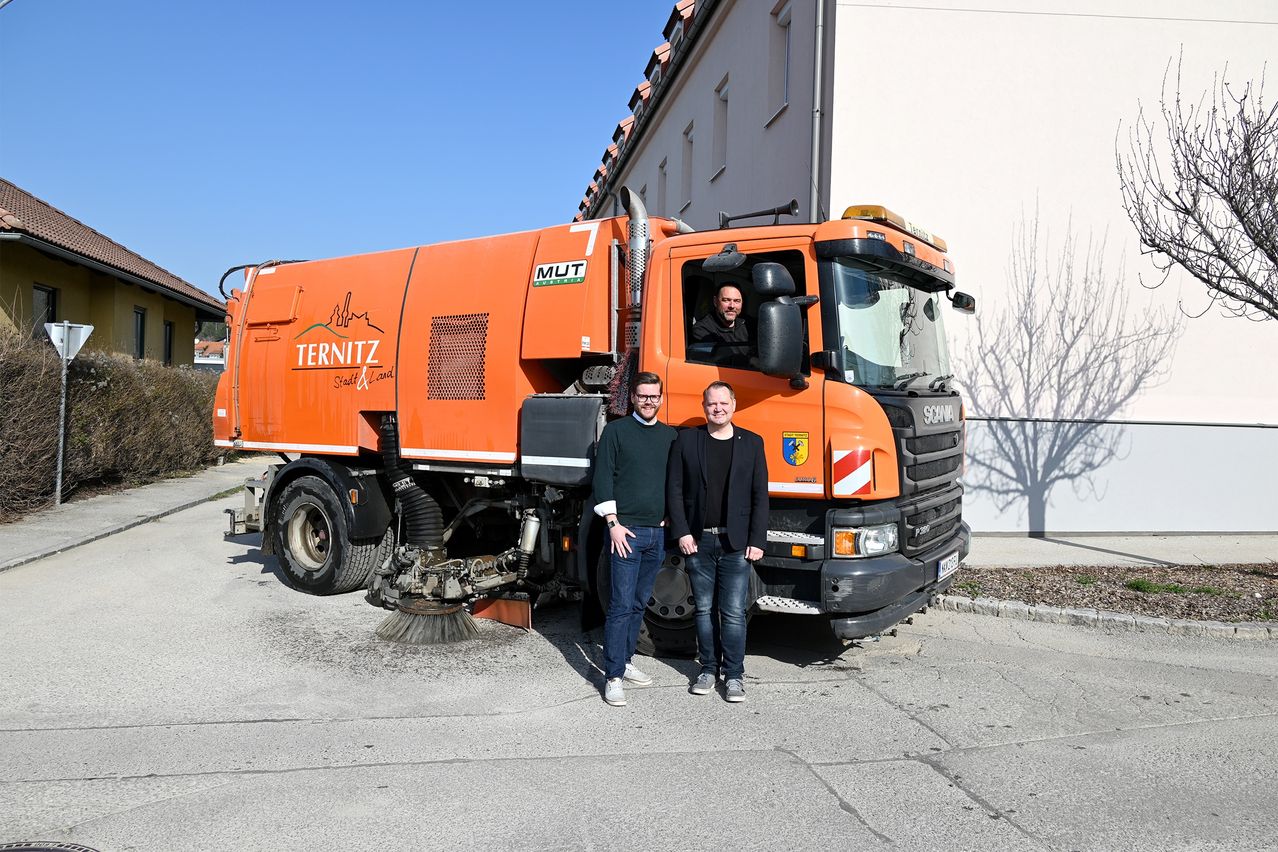 Zwei Männer stehen vor einem orangefarbenen Müllwagen mit der Aufschrift Ternitz, wobei ein Mann auf dem Fahrersitz sitzt.