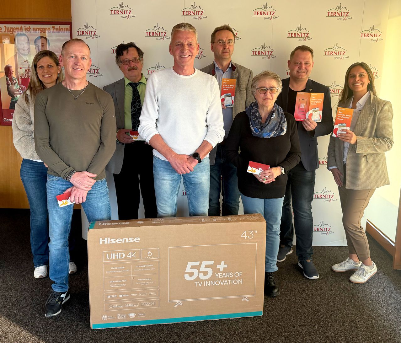Eine Gruppe von Menschen steht hinter einem großen Hisense UHD 4K Fernseher. Sie halten Broschüren und lächeln. Sie sind alle formal gekleidet.