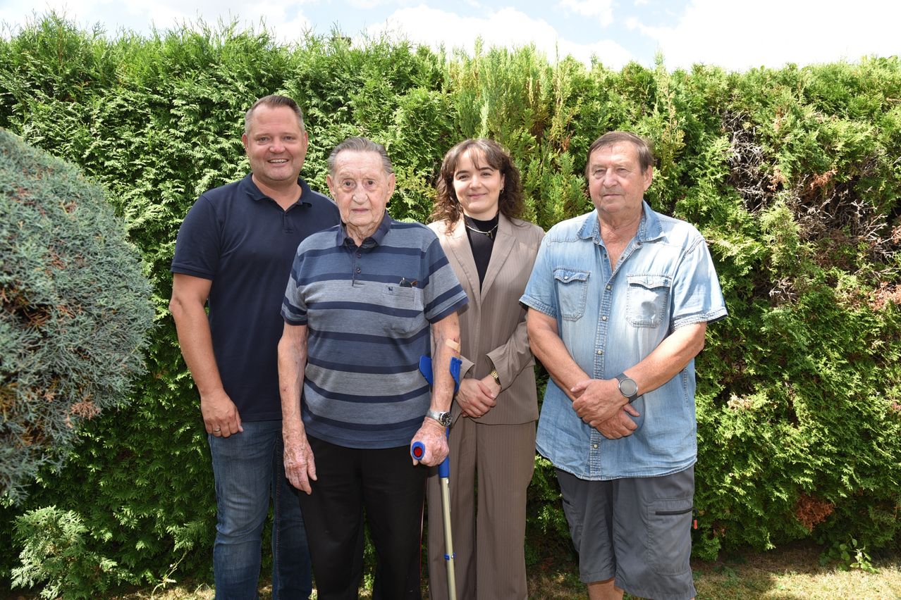 Four individuals stand together in front of a hedge, smiling for a photo. An elderly man uses a cane, while the others have their hands on their hips.