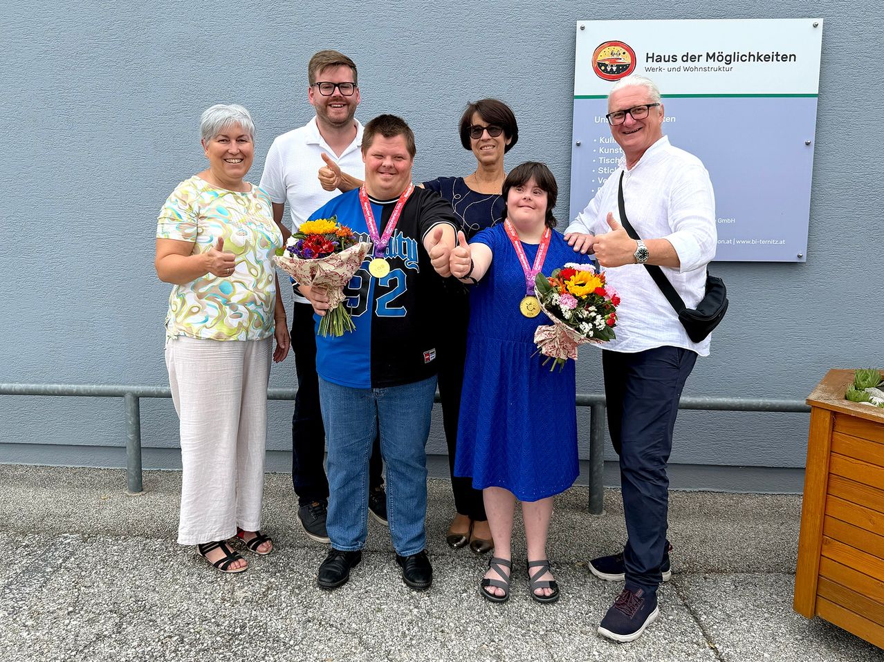 A group of people, including two individuals with disabilities, are posing for a photo with flowers and medals. They are standing in front of a sign that reads 'Haus der Möglichkeiten'.