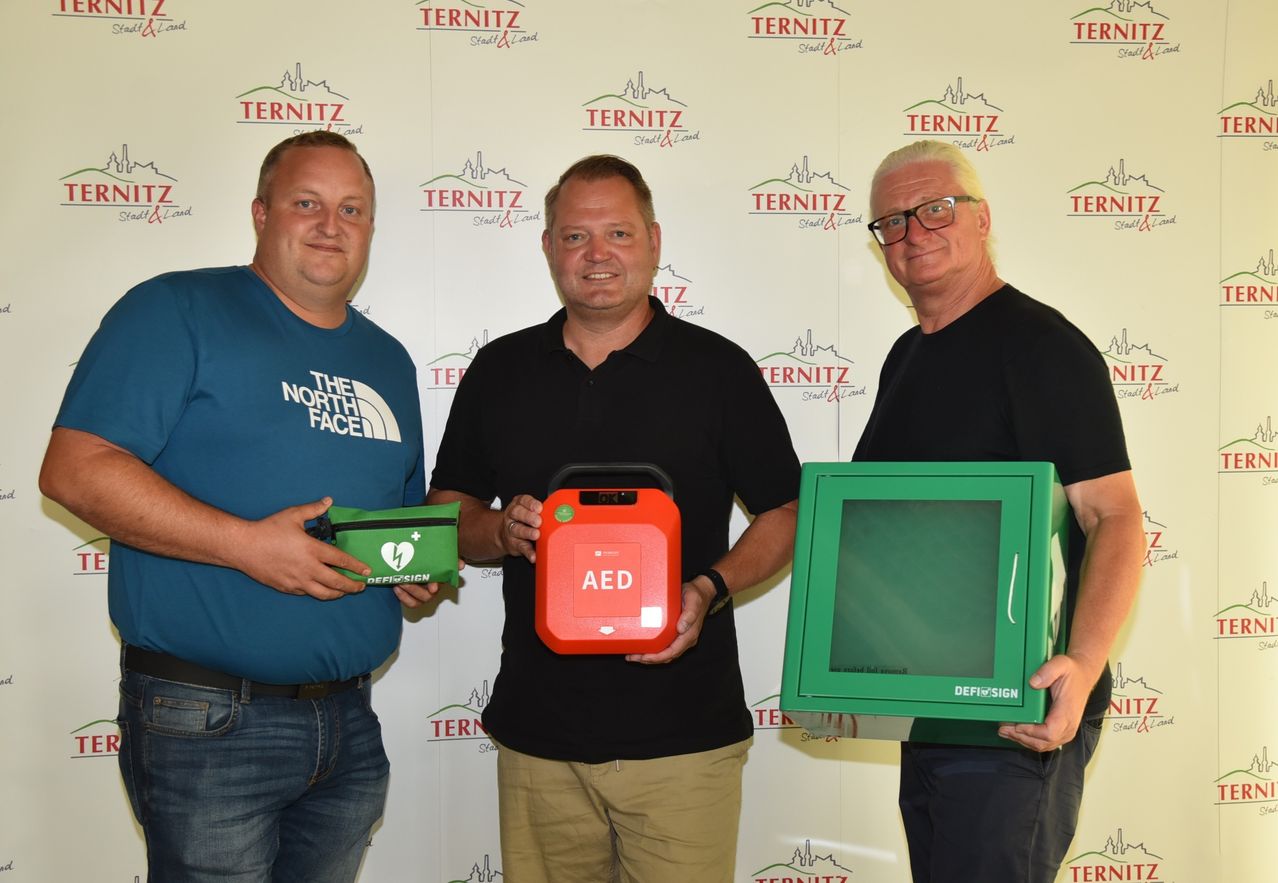 Drei Männer stehen vor einer Wand mit Logos und halten eine grüne Tasche, ein rotes AED-Gerät und eine grüne DEFISIGN-Box.
