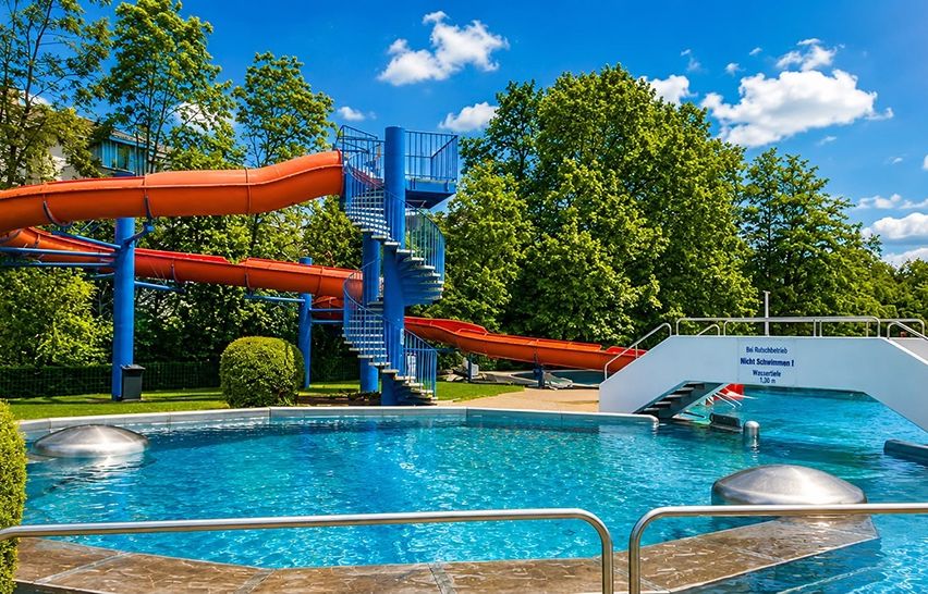 Ein Freibad mit einer großen blauen und orangefarbenen Rutsche, einer Wendeltreppe und einem Schwimmbecken unter blauem Himmel mit Wolken.