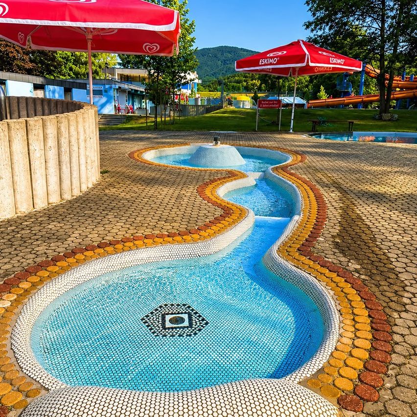 Ein Wasserspiel mit Mosaikdesign in einem Park, umgeben von roten Sonnenschirmen und einem Rasenbereich mit einem Gebäude und Wasserrutschen im Hintergrund.