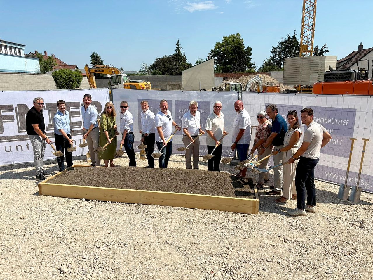 Eine Gruppe von Menschen in formeller Kleidung versammelt sich um eine mit Kies gefüllte Holzkiste, hält Schaufeln und posiert für ein Foto an einer Baustelle.