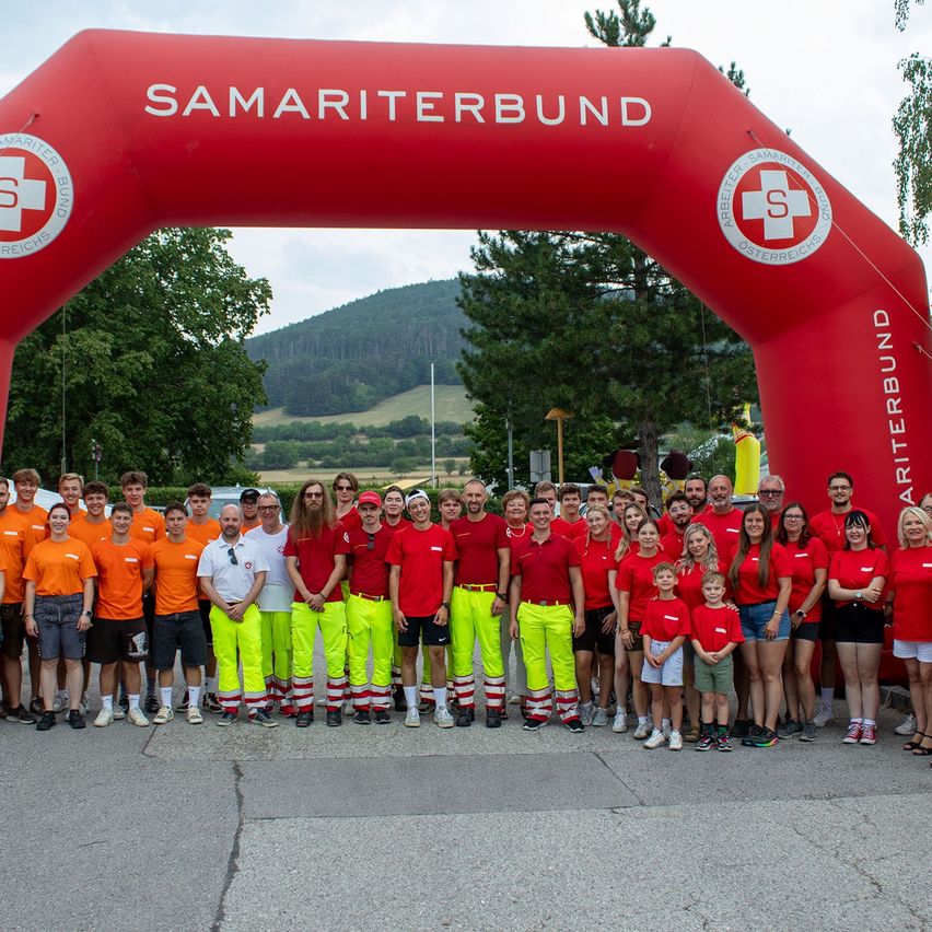 A group of people in red and yellow uniforms, possibly volunteers, are posing for a photo in front of a red arch with the words 'Samariterbund'.