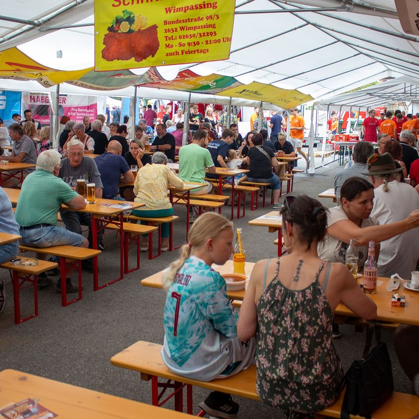 A crowded outdoor dining area with tents, people sitting at wooden benches, and a sign advertising 'Schrimps Wimppassig'.