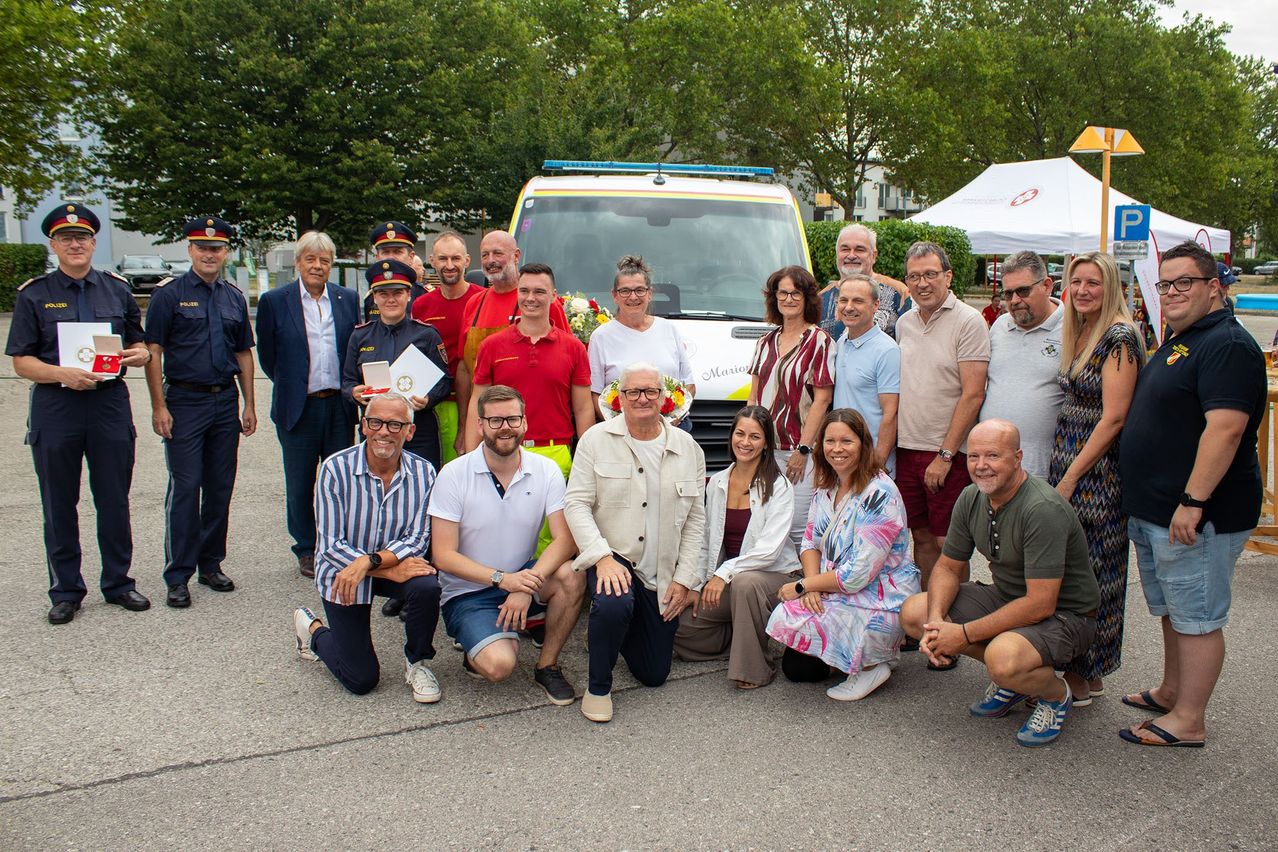 A group of people in various uniforms and casual attire are posing for a photo in front of an ambulance. Some hold flowers and awards, and the background features trees and a tent.