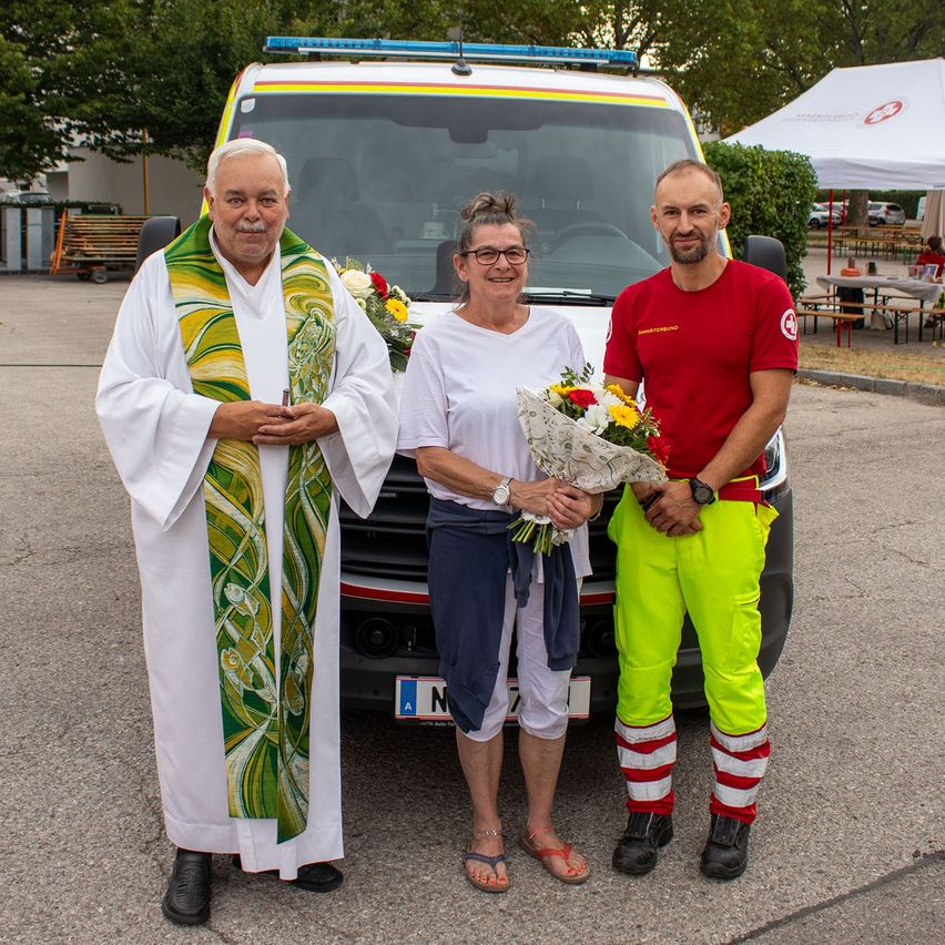 Three individuals stand in front of an ambulance, with a priest on the left, a woman with flowers in the middle, and a man in a red shirt on the right.