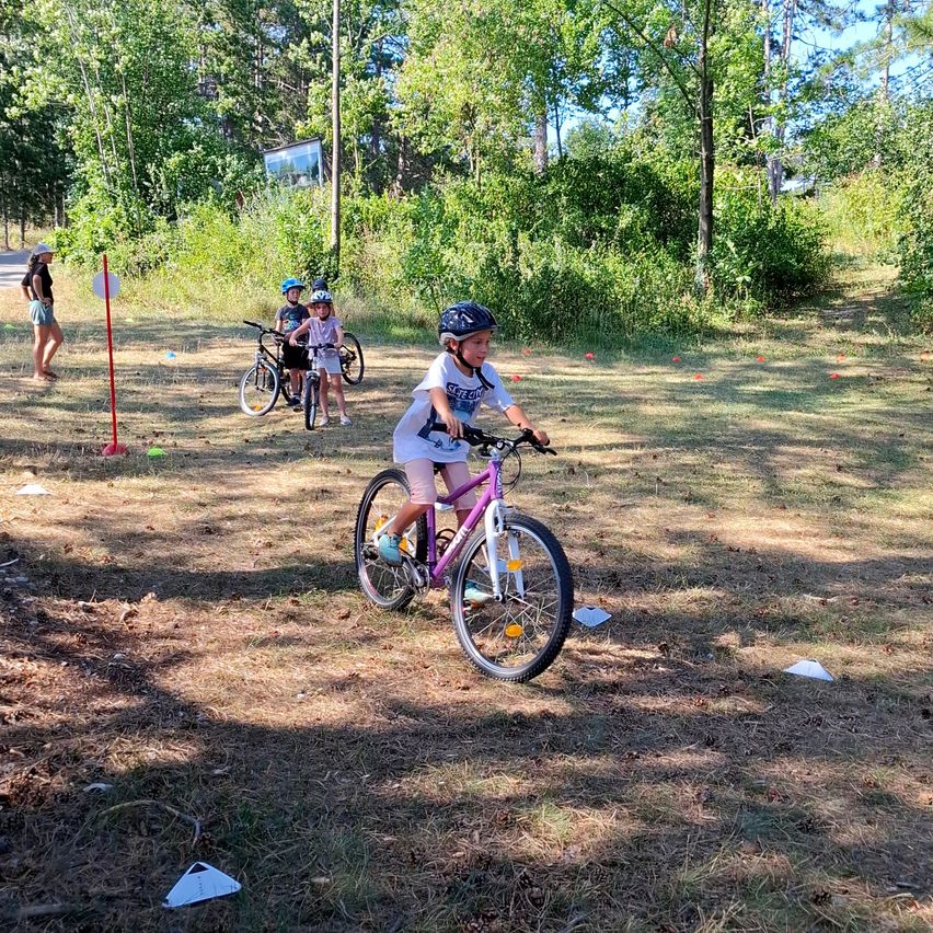 A young girl rides a purple bicycle on a dirt path, surrounded by other children and adults. They are all wearing helmets and appear to be participating in a bike riding activity.