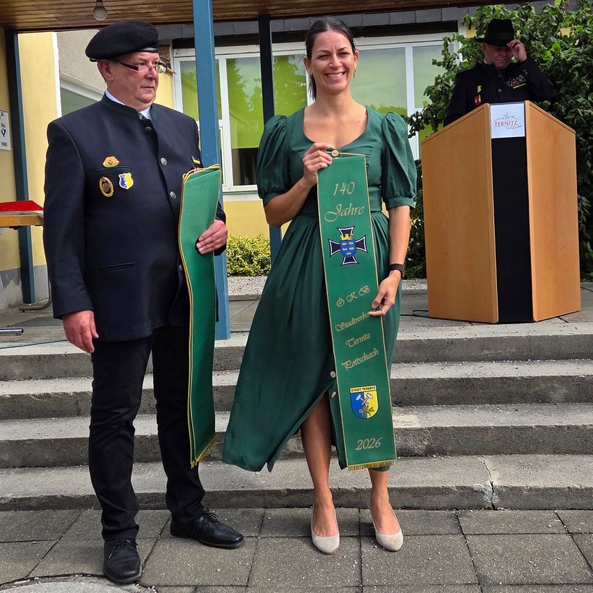 A man and a woman stand on steps, the woman holding a green banner with '140 Jahre' and '2026' written on it, smiling and posing for a photo. Behind them, a podium with a sign reading 'TERNITZ' is visible.