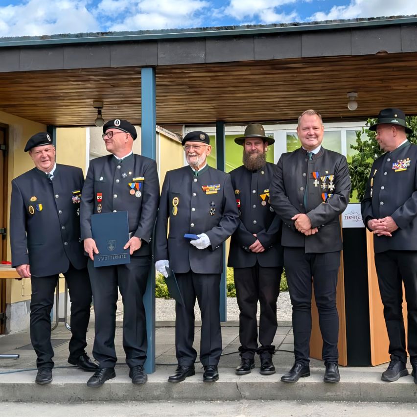 Six men in uniform stand on a porch, smiling for a photo. They are adorned with medals and one holds a blue folder. Behind them, a wooden structure with a sign is visible.