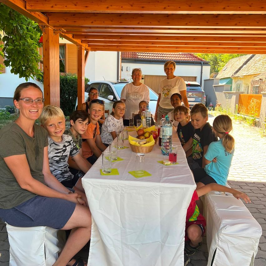 A group of people, including children, are seated around a table in a backyard. The table is covered with a white tablecloth and has various items such as fruits, bottles, and glasses. The setting is outdoors under a wooden pergola.