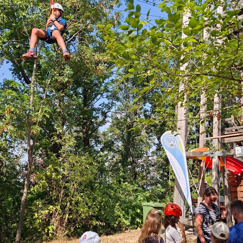 A child is ziplining through a forest, wearing a helmet and harness. Several people are watching from below, including a woman with a striped shirt and a child in a red helmet.