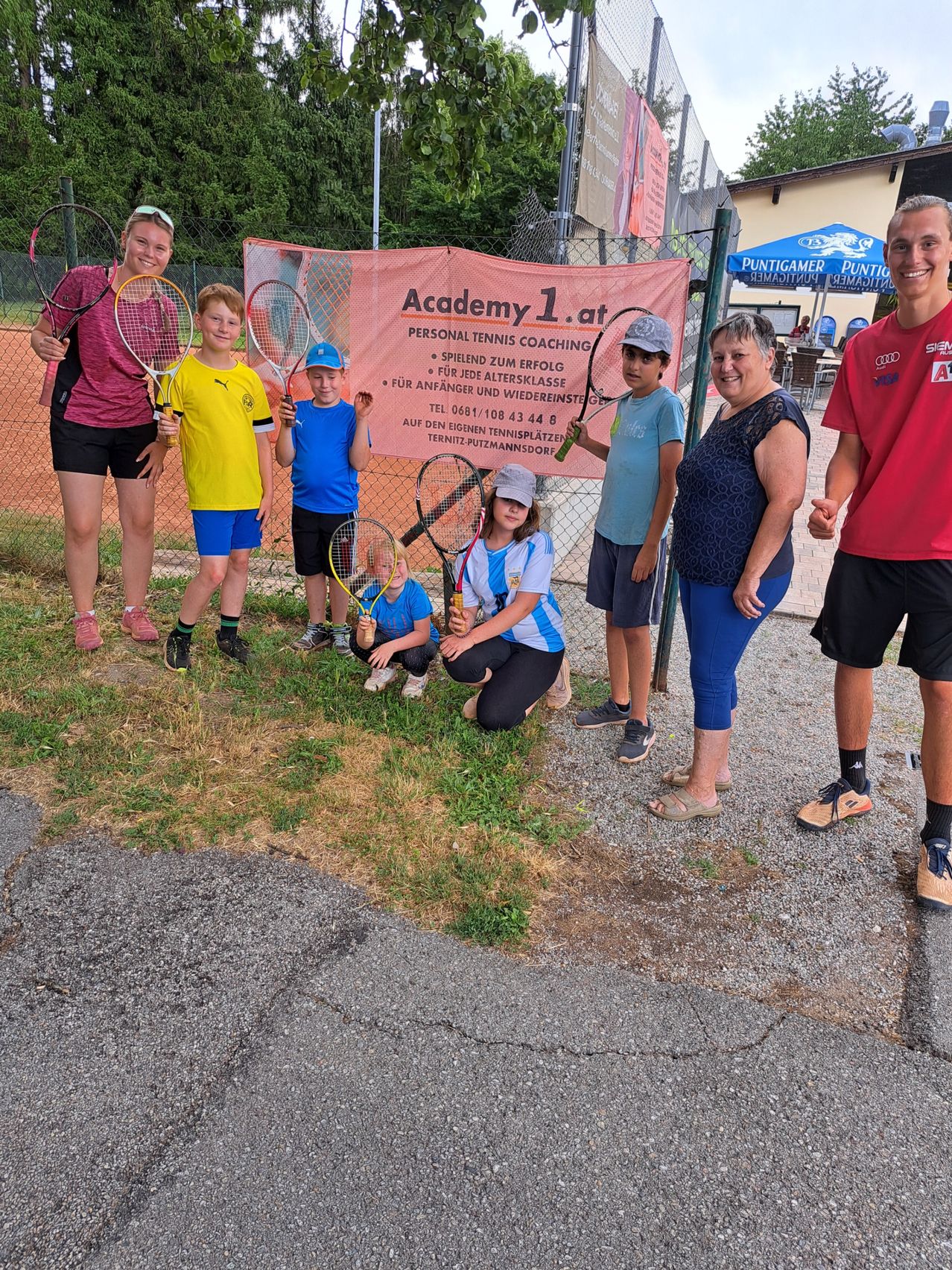 A group of children and adults are posing for a photo at a tennis court with a banner that reads Academy 1.at.