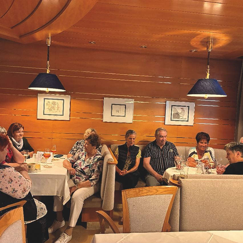 A group of elderly people are sitting at a table in a restaurant, with some of them drinking wine. The restaurant has wooden walls and ceiling, and there are three picture frames hanging on the wall.