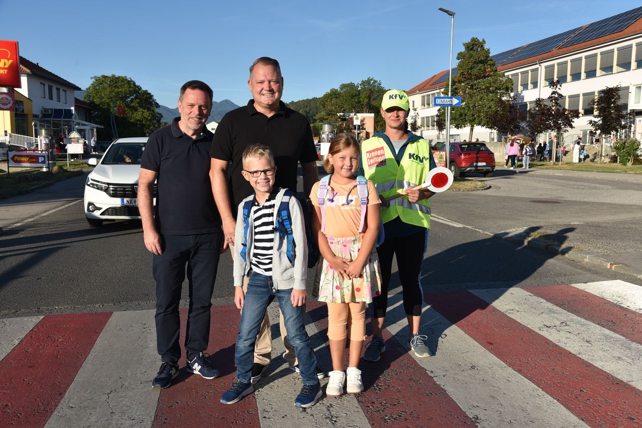 A group of people, including two adults and two children, are standing on a pedestrian crossing. The adults are smiling, and one is wearing a reflective vest. The children are wearing backpacks. Behind them, there is a building with solar panels on the roof.
