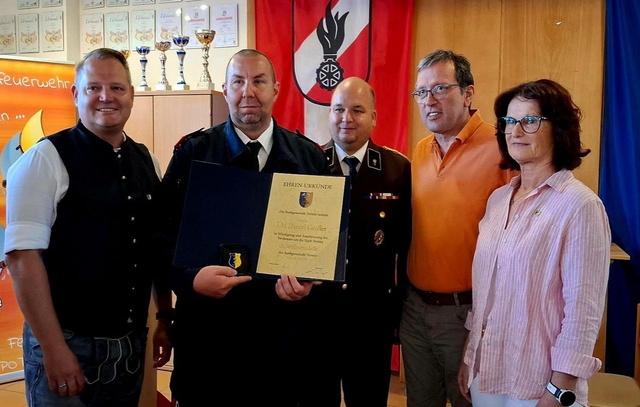 A man in uniform holds a certificate, flanked by three others in a room with a flag and trophies in the background.