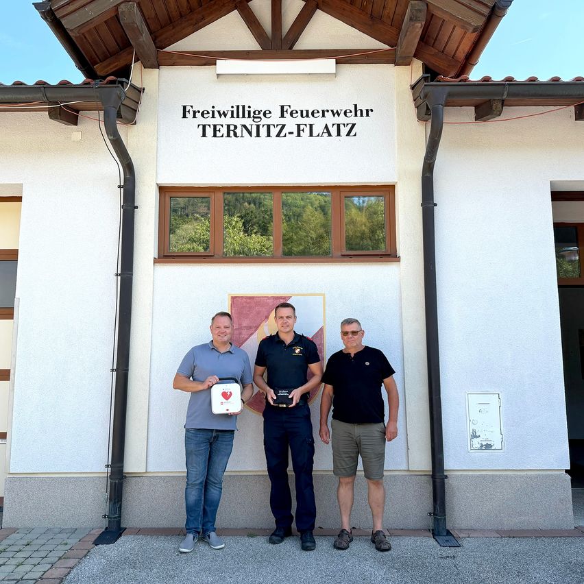 Three men stand in front of a building with a sign reading 'Freiwillige Feuerwehr TERNITZ-FLATZ'. The man in the middle holds a trophy, and the man on the left holds a box.