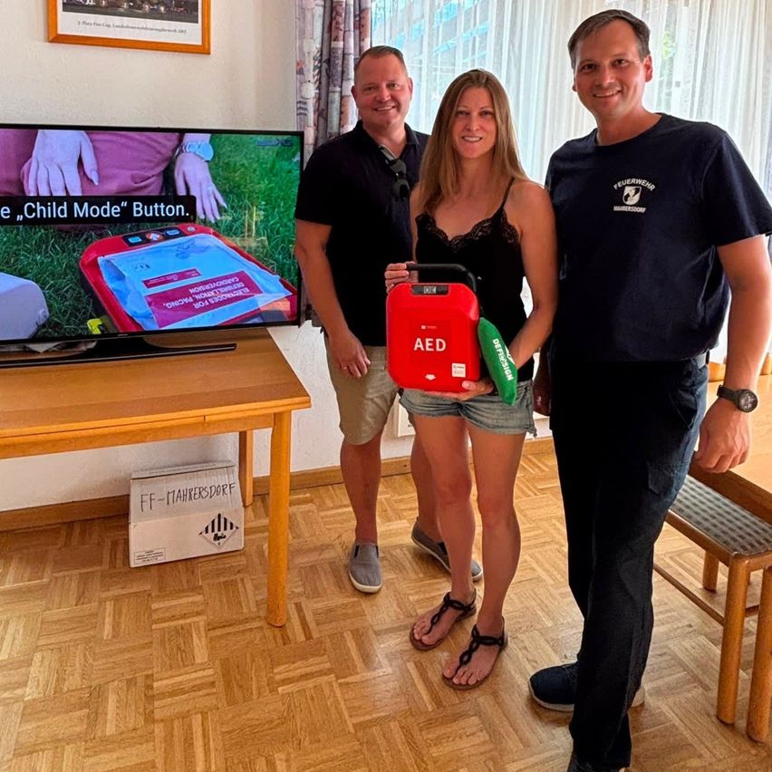 Three people stand in a room, one holding an AED device, with a TV screen behind them showing a safety message.