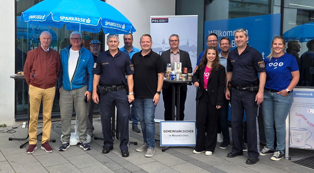 Eine Gruppe von Personen, darunter Polizeibeamte und eine Frau, posiert für ein Foto an einem Polizeistand mit einem Banner, das 'Gemeinsam.Sicher in Neunkirchen' besagt.