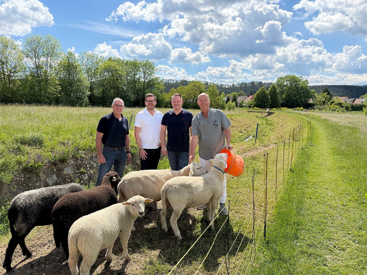 Vier Männer stehen in einem Feld mit Schafen, einer hält einen Eimer. Hinter ihnen umgibt ein Zaun einen grasbewachsenen Bereich. Bäume und Häuser im Hintergrund unter einem teilweise bewölkten Himmel.