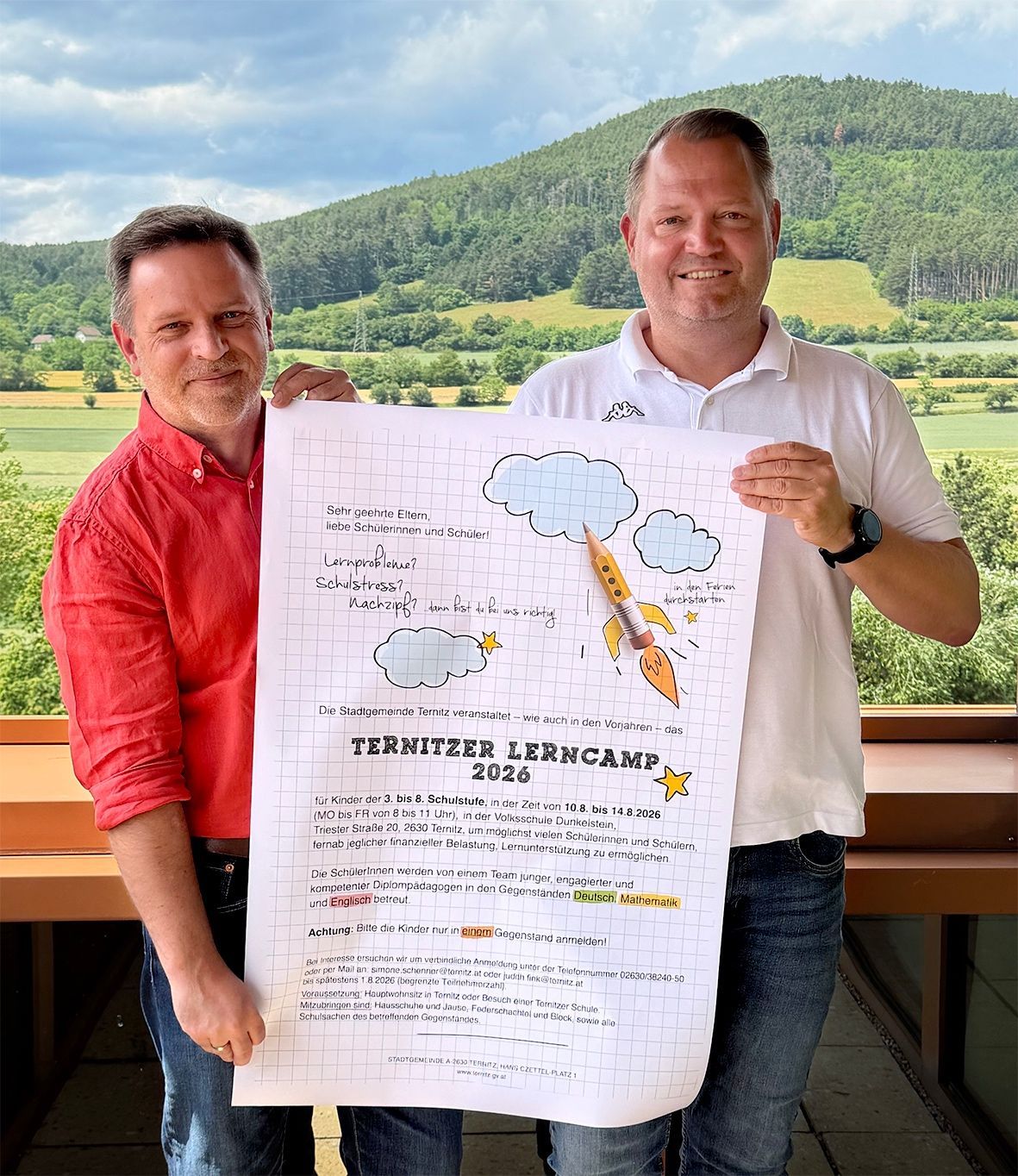 Zwei Männer stehen in einer malerischen Außenumgebung und halten ein großes Plakat. Das Plakat hat deutschen Text und eine Karikatur einer Rakete. Ein Mann trägt ein rotes Hemd, der andere ein weißes Poloshirt. Hinter ihnen ist eine Aussicht auf ein Tal mit Bergen und Grün.