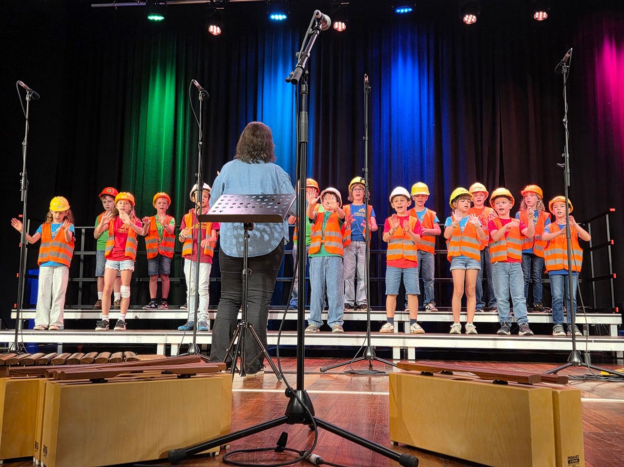Ein Kinderchor in Sicherheitswesten tritt mit einer Frau vor einem Mikrofon und Notenständer auf der Bühne auf.