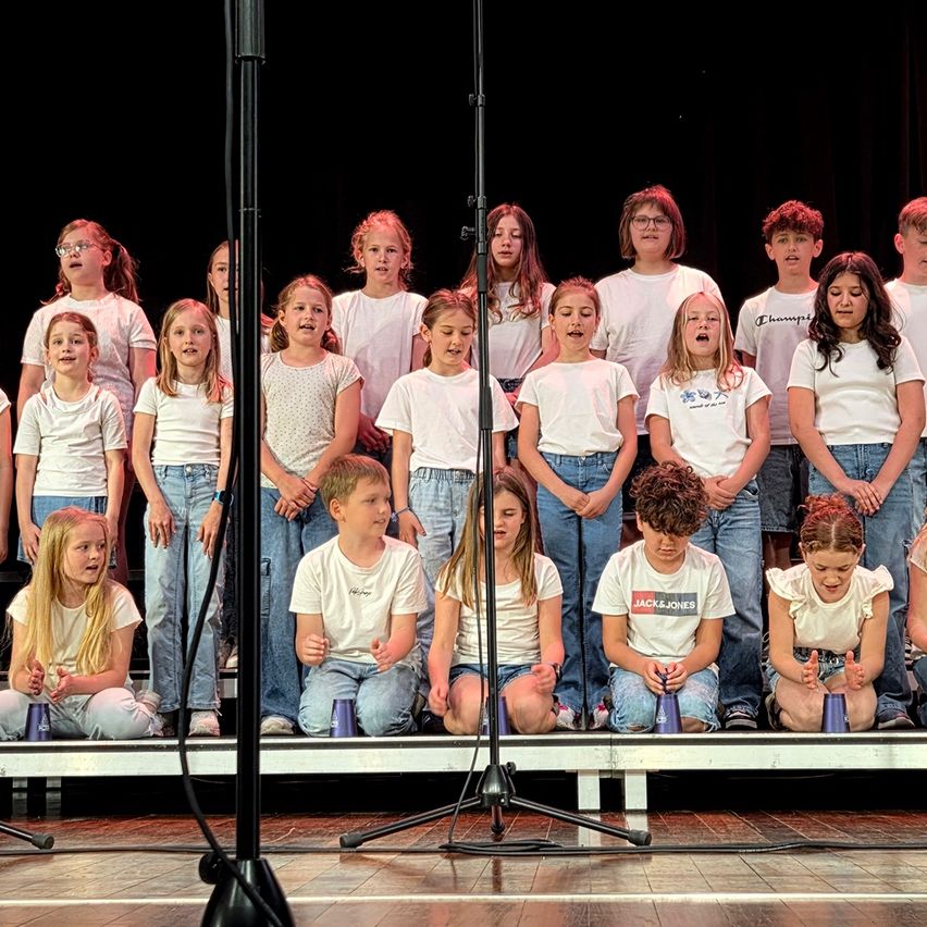 Ein Kinderchor auf der Bühne, in weißen T-Shirts gekleidet, einige kniend und andere stehend, performt vor einem schwarzen Hintergrund.