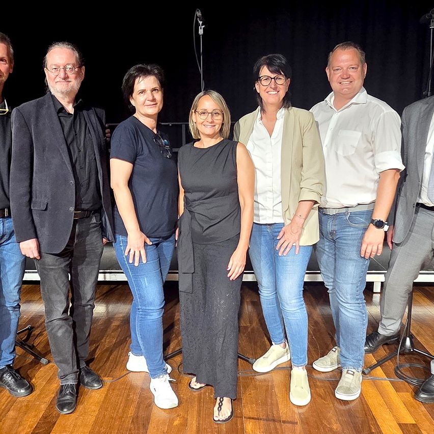 Eine Gruppe von sechs Personen, drei Männer und drei Frauen, stehen gemeinsam auf einem Holzboden und lächeln für ein Foto. Sie sind lässig gekleidet und scheinen ein Team zu sein.