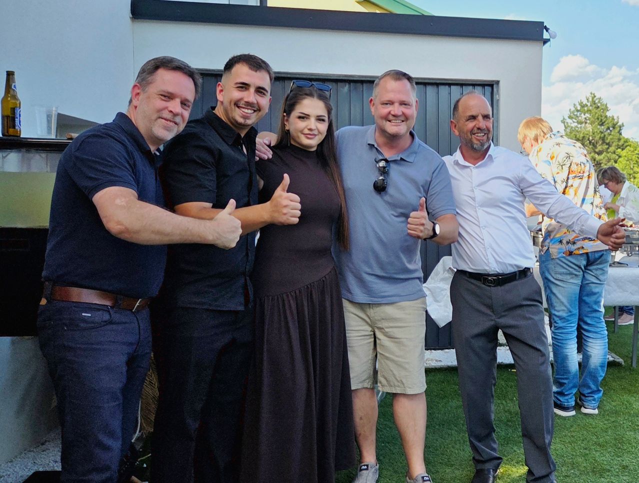 Fünf Männer und eine Frau stehen zusammen und lächeln für ein Foto auf einem grünen Rasen. Sie alle zeigen Daumen hoch. Hinter ihnen ein Gebäude mit einer Metallwand und einem klaren Himmel.