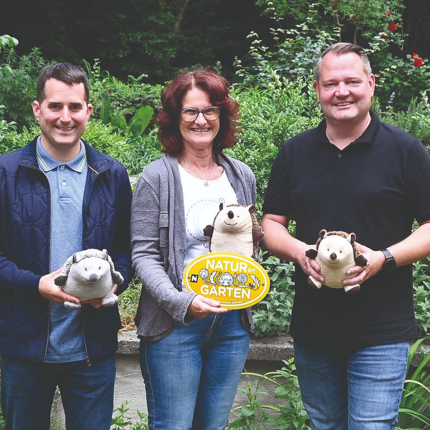 Three people stand in a garden, holding stuffed animals. A man on the right smiles, while the man on the left looks at the camera. The woman in the middle holds a yellow sign with the words 'Natur im Garten'. Behind them are various plants and flowers.
