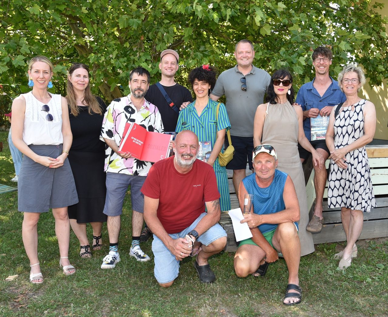 A group of people, including men and women, stand and crouch on grass under trees. Some hold books and glasses. They all smile for a photo.