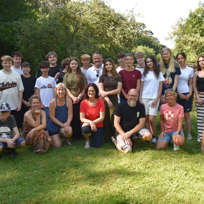 A group photo of adults and children on a sunny day, with a grassy area and trees in the background. They are all smiling and posing for the camera.