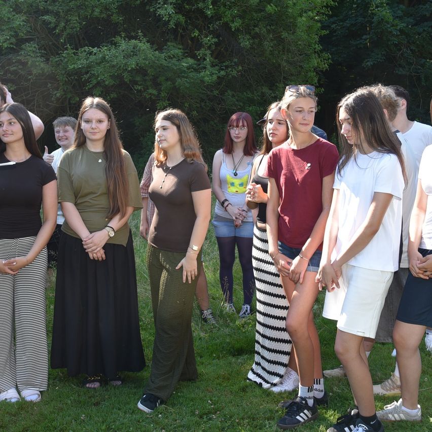 A group of young people are standing in a grassy area, possibly for a photo. They are all looking in the same direction. Some of them are wearing bracelets and necklaces. The background is blurry, with trees and a forest.