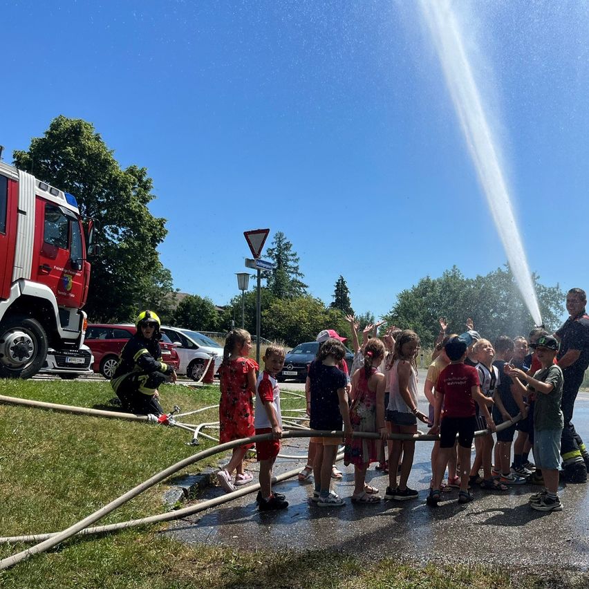 A group of children stands in line holding a fire hose while a firefighter sprays water from the nozzle. A fire truck and several cars are parked nearby.