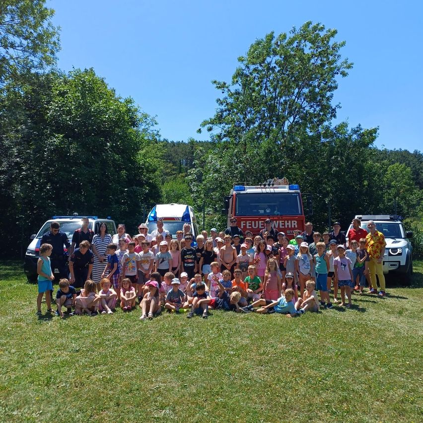A group of children and adults pose for a photo in a grassy field with fire trucks and vehicles in the background.