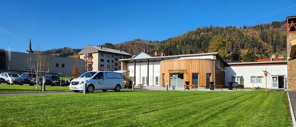 Ein weißer Van ist vor einem modernen Gebäude mit einem grünen Rasen und Bergen im Hintergrund geparkt.