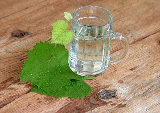 Ein Glaskrug mit klarem Wasser steht auf einer Holzoberfläche, daneben einige grüne Blätter und Wassertropfen.