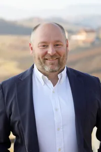 Ein Mann mit Bart trägt einen dunkelblauen Blazer und ein weißes Hemd. Er steht draußen und lächelt in die Kamera.